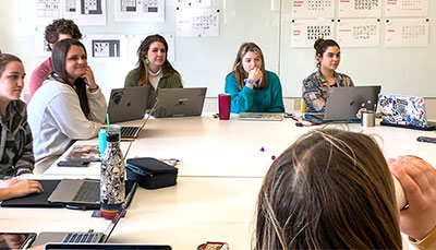 Group of students sitting around a table with projects pinned to a board behind them.